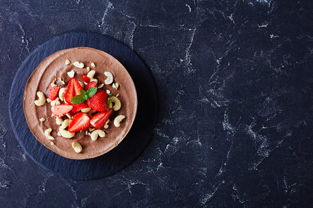 Plant based, no bake vegan dessert chocolate cheesecake with no-bake crust of dates, cocoa, and pecans with strawberries and mint on top on a dark concrete background, top view, copy spaceの写真素材