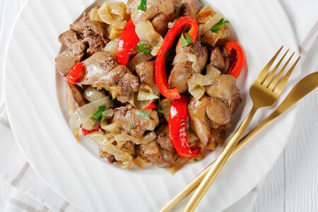 High protein appetizer: fried chicken liver with onion rings and red bell pepper, fresh parsley on top on a white wooden background with golden cutlery and napkin, top view, close-upの写真素材