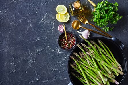 fresh green asparagus in black baking dish with golden cutlery, lemon slices, peppercorns and bottle of olive oil on grey concrete table, horizontal view from above, flat lay, free spaceの写真素材