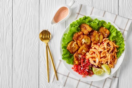 Latin american dish: fried plantains served with shredded chicken mixed with buffalo sauce, pico de gallo salsa, lime, lettuce leaves, on a white plate on a wooden background, flat lay, copy spaceの写真素材