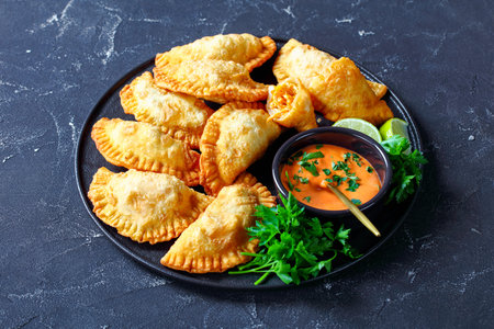deep-fried Buffalo Chicken Empanadas with Low-Calorie Dip on a black platter on a concrete table,...