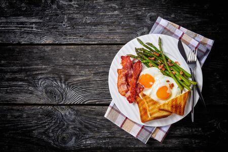fried sunny side up eggs, crispy bacon, toasts, asparagus on a white plate with silver fork and knife, on an old wooden table, flat lay, free space, horizontal view from aboveの写真素材