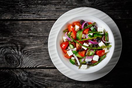 Greek salad with grilled asparagus, cherry tomatoes, cucumbers, black olives, red onion rings with olive oil, soft feta cheese served on a  plate on a wooden table, view from above, copy spaceの写真素材
