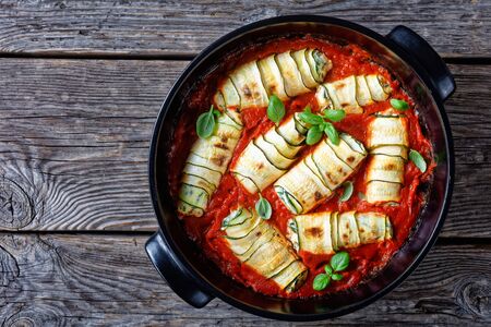 Zucchini roll-ups stuffed with fresh italian ricotta with spinach and basil leaves, garlic baked in tomato sauce, served on a black baking dish on rustic wooden background, top view, copy spaceの写真素材