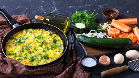 Healthy breakfast: vegetable and chicken eggs frittata of shredded  zucchini and carrot served on a frying pan on a dark concrete background with olive oil, parsley, ingredients, close-upの写真素材