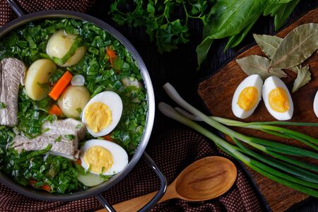 Summer greens sorrel borscht or soup of fresh sorrel, green onion with pork ribs, young potato, carrot, and boiled eggs, served in a  stock pot on a wooden background with a wooden spoon, top viewの写真素材
