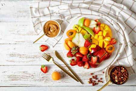 Colorful fruit dessert: platter of strawberries, mango, apples, pears, apricots, with peanut butter and peanuts on a white plate on a white wooden table, top view, flat lay, copy spaceの写真素材