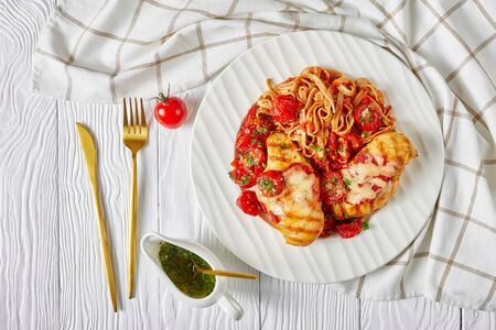 Grilled chicken breasts with melted mozzarella cheese on top, whole wheat linguine, cherry tomatoes, and garlic herb sauce, golden cutlery on a white wooden background, flat layの写真素材