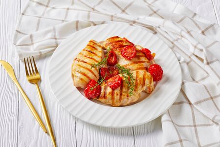 Grilled chicken breasts with roasted cherry tomatoes on a white plate on a wooden table with kitchen towelの写真素材