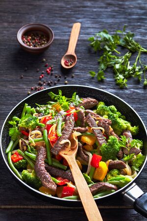 Colorful fajitas of beef steak with broccoli, green bean, yellow and red sweet peppers, parsley, onion roasted on a skillet with peppercorns on a dark wooden background, close-upの写真素材