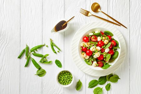 Summer salad of mini mozzarella balls, fresh baby spinach cherry tomatoes and young peas with balsamic vinegar dressing on a white plate on a white wooden background, top view, flat layの写真素材