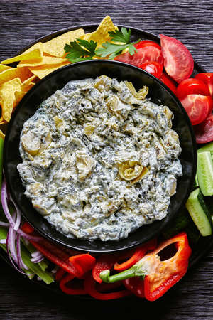 Spinach Artichoke leafy cheese Dip in a bowl with Tortilla Chips, cucumber, celery sticks, tomato slices and red bell pepper, vertical view from above, flat layの写真素材