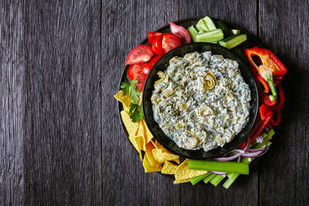 Spinach Artichoke leafy cheese Dip in a bowl with Tortilla Chips, cucumber, celery sticks, tomato slices and red bell pepper, horizontal view from above, flat lay, free spaceの写真素材