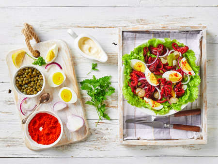 baby octopus salad with hard-boiled eggs, capers, marinated onion on lettuce leaves in a bowl on a tray, on a rustic wooden table with ingredients, flat layの写真素材