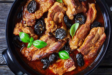 Stewed short pork ribs with dried plums or prunes, pork broth, onion, and garlic, decorated with fresh basil, served on a black baking dish on an old barn wood background, close-upの写真素材