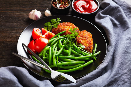 close-up cornmeal-crusted minced meat patties served with blanched green beans and fresh tomatoes on...