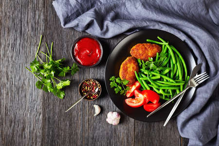 cornmeal-crusted minced meat patties served with steamed green beans and fresh tomatoes on a black plate on a dark wooden table, horizontal view from above, flat lay, free spaceの写真素材