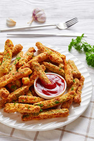 close-up of crunchy zucchini sticks breaded with panko breadcrumbs, parmesan cheese, spices on a white plate with ketchup on a wooden table, vertical viewの写真素材