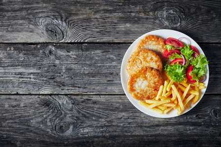 fried turkey burgers served with lettuce tomato salad and french fries on a white plate on a rustic wooden table, horizontal view from above, flatlay, empty spaceの写真素材