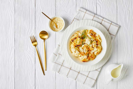 Cauliflower cheese with garlic and thyme served on a white plate with golden cutlery on white wooden background with a napkin, shredded cheddar cheese, and milk, top view, flat layの写真素材