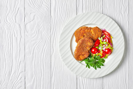breaded lamb chops served with tomato, red onion, green pepper salad on a white plate on a wooden...