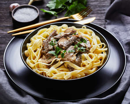 beef stroganoff cooked with sour cream mushrooms sauce, served with egg noodles in a black bowl with golden fork and knife on a dark wooden table, close-up, landscape viewの写真素材