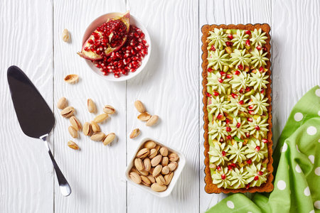 Pistachio ganache tart with pomegranate seeds on a square baking tin made of shortcrust pastry and whipped white chocolate pistachio filling served on a white wooden background with dessert paddleの写真素材