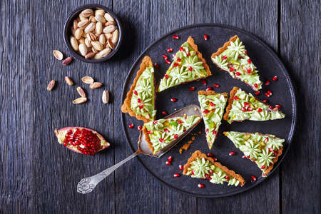 Pistachio ganache tart with pomegranate jelly, pomegranate seeds on top, served on a black plate served on a dark rustic wooden table with dessert paddle and linen dish towel, top view, close-upの写真素材