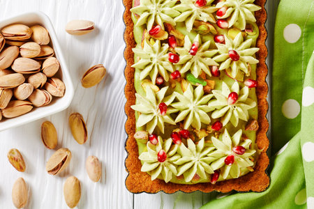 Pistachio ganache tart with pomegranate seeds on a square baking tin made of shortcrust pastry and whipped white chocolate pistachio filling served on a white wooden table, top view, close-upの写真素材