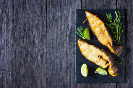 Traditional smoked halibut fish steaks with lime and rosemary sprigs on a black slate on a dark...