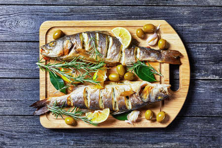 Whole roasted seabass fish with lemon wedges and herbs: rosemary, parsley, and bay leaf, served on a wooden cutting board with green olives and tartar sauce, top view, close-upの写真素材