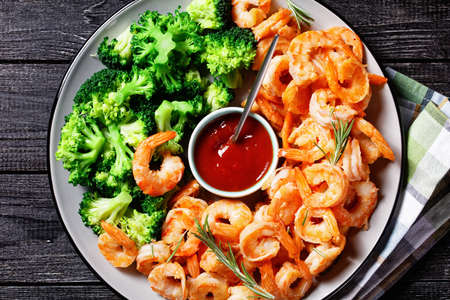 Garlic butter prawns or shrimps deshelled and fried served with steamed broccoli, ketchup, and lime on a plate with rosemary sprigs and cutlery on a dark wooden background, top view, close-upの写真素材