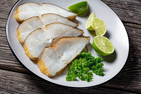 cold-smoked halibut fish slices served with lime on a plate on a rustic wooden table, horizontal...