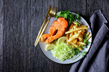 salmon steaks served with potato fries and coleslaw on a plate on a dark wooden table, horizontal view from above, flat lay, free spaceの写真素材