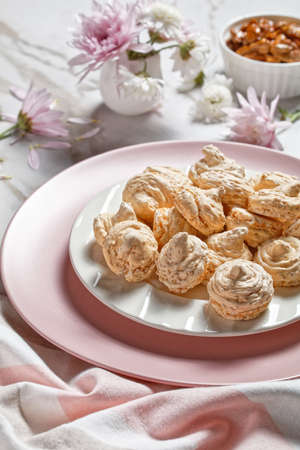 Swirled meringue cookies with caramelized walnuts of egg whites whipped with sugar served on a white plate on a light marble table with flowers, top view, close-upの写真素材