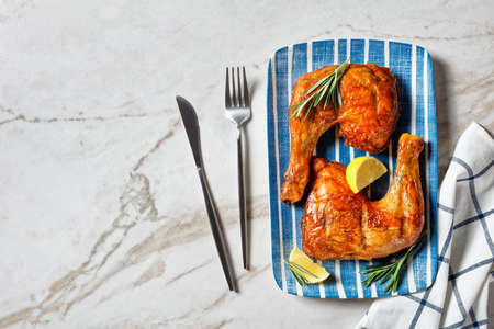 Golden brown chicken leg quarters on a plate with cutlery, lemon wedges, and fresh rosemary sprigs on a white marble stone background, top view, copy spaceの写真素材