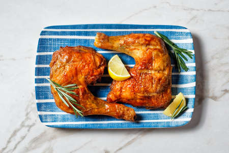 Chicken leg quarters on a plate with cutlery, lemon wedges, and fresh rosemary sprigs on a white marble stone background, top view, close-upの写真素材