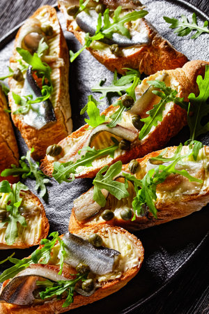anchovy butter sandwiches with fresh rocket salad leaves, capers on a black platter on a rustic wooden table, vertical view from aboveの写真素材