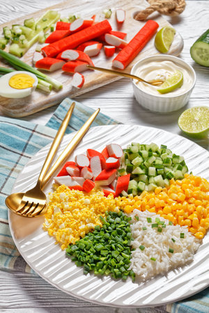 Crab salad of surimi sticks with canned corn, cucumbers, spring onion hard-boiled eggs, jasmine rice divided into sectors on a white plate on a white wooden background, top view, close-upの写真素材