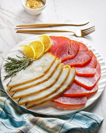Smoked fish platter: cold smoked halibut and cold smoked tuna slices served on a white plate with rosemary sprigs, lemon wedges and English mustard on a light marble background, top view, close-upの写真素材