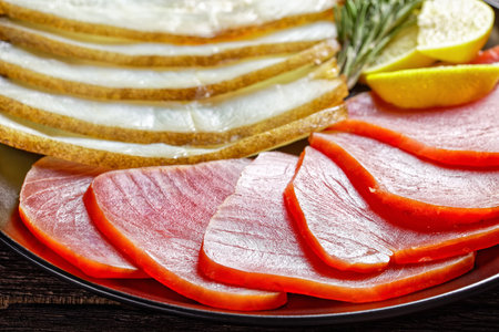Smoked fish appetizer plate: cold smoked halibut and cold smoked tuna slices served on a black plate with rosemary sprigs and lemon wedges on a dark wooden table, top view, close-upの写真素材