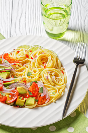 linguine with avocado, tomatoes and lime sprinkled with chopped chili and red onion on a white plate on a wooden table served with a glass of water, vertical view from above, close-upの写真素材