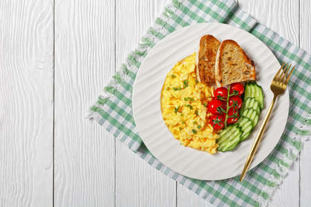 scrambled eggs with sliced cucumber, tomatoes, toasted bread and a glass of fresh orange juice on a white plate on a wooden textured table, flat lay, free spaceの写真素材
