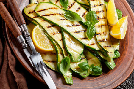 grilled zucchini slices on a clay plate with lemon, olive oil, spices and fresh basil leaves on a dark wooden table, italian cuisineの写真素材