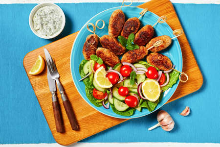 Lamb kofta kebabs of ground lamb, served on a blue plate with yogurt dressing, fresh vegetables salad, and lemon wedges, greek cuisine on a white wooden table, top view, close-upの写真素材
