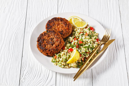 harissa lamb burgers served with pearl couscous salad on a plate with lemon slices on a white wooden...