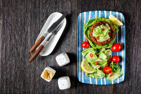 open sandwich with lamb burger on a toasted bun with cucumber ribbons and tomatoes salad on a plate on a dark wooden table, horizontal view from aboveの写真素材