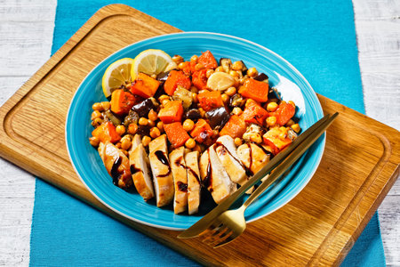 Moroccan salad of roasted vegetables: pumpkin, eggplant, with chickpeas, lemon and baked chicken breast, served on a plate with cutlery on a white wooden background, top view, close-upの写真素材