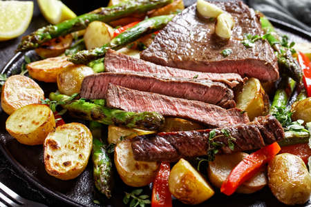 close-up of sliced beef steak with roasted vegetables new baby potatoes, asparagus and red pepper on a black plateの写真素材