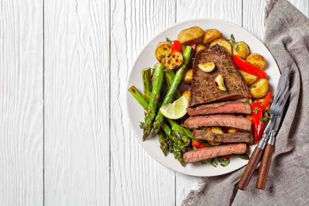 beef steak with barbequed new baby potatoes, asparagus, lemon and red pepper on a white plate with barbecue sauce on a wooden table, flat lay, free spaceの写真素材